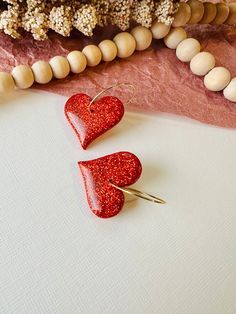 two red heart shaped earrings sitting on top of a white surface next to beads and flowers