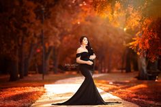 a pregnant woman in a black gown standing on a path surrounded by trees and leaves