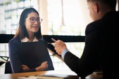 a woman sitting at a desk talking to a man who is holding a clipboard