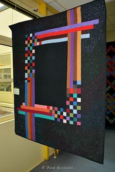 there is a large black quilt hanging from the ceiling in front of a window with colorful squares on it