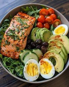 a salad with salmon, avocado, tomatoes and olives in it on a wooden table