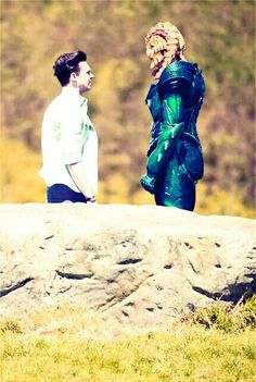 two people standing next to each other on top of a rock near a grass covered field