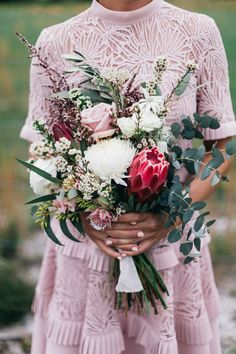 a woman holding a bouquet of flowers and greenery in her hands while standing outside