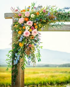 a wooden cross with flowers and greenery hanging from it's sides in front of a grassy field