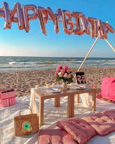 a table on the beach with pink pillows and balloons that say happy birthday above it