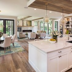 an open concept kitchen and living room in a house with hardwood floors, large windows, white cabinets, and wooden ceiling