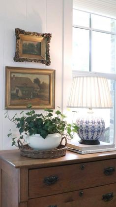 a wooden dresser topped with a potted plant next to a lamp and framed pictures