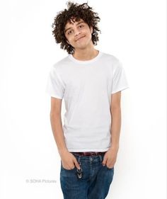 a young man with curly hair standing in front of a white wall wearing jeans and a t - shirt