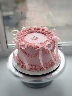 a pink cake sitting on top of a silver plate next to a window with white ribbons