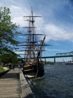 an old sailing ship is docked at the end of a wooden pier with people walking on it