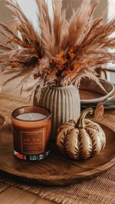 a candle and some pumpkins on a wooden plate with flowers in the vase next to it