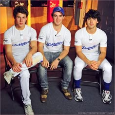 three young men sitting next to each other wearing baseball caps and white shirts with blue lettering on them