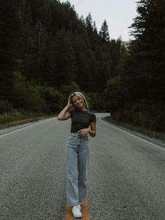 a woman standing on the side of a road with her hands behind her head and trees in the background