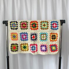 a crocheted blanket is hanging on a black stand with white curtains in the background