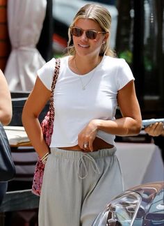 a woman in white shirt and grey pants standing next to a car with sunglasses on