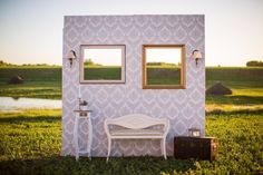 a white bench sitting in the grass next to a wall with two mirrors on it