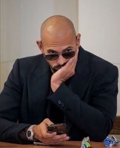 a bald man wearing sunglasses and holding his hand to his face while sitting at a table