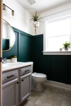 a white toilet sitting next to a bathroom sink in a green painted room with potted plants on the window sill