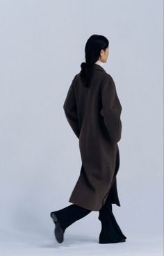 a woman walking across a white floor wearing a brown coat and black pants with her back turned to the camera