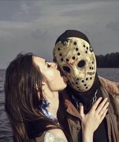 a woman kissing a man wearing a mask in front of the water on a cloudy day