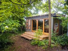 a small wooden cabin in the woods surrounded by trees and bushes, with an outside deck