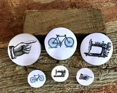 four buttons with hand drawn images of sewing machines and bicycles on them, sitting on a piece of wood