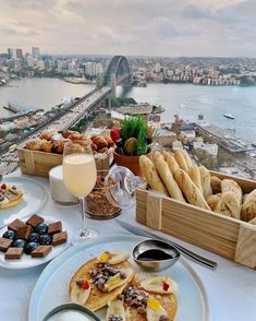 a table topped with plates of food next to a large body of water and a bridge in the background