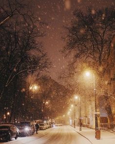 a snowy street at night with cars parked on the side and people walking down the sidewalk