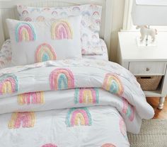 a white bed with rainbows and clouds on it's comforter, next to a night stand