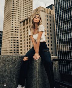 a woman sitting on top of a cement wall in front of tall buildings with her legs crossed