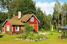 a small red house in the middle of a green yard with trees and bushes surrounding it