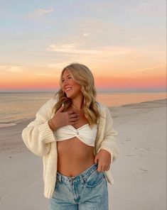 a woman standing on top of a sandy beach next to the ocean wearing jeans and a sweater