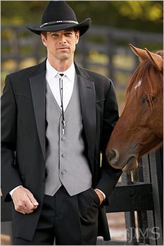 a man standing next to a brown horse wearing a black hat and suit with his hands in his pockets