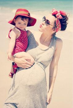 a woman holding a baby girl on the beach while wearing a red hat and sunglasses