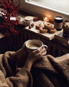 a person wrapped in a blanket holding a cup of coffee with donuts on the side