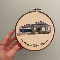 a hand holding up a embroidery hoop with a house in the middle and trees on it