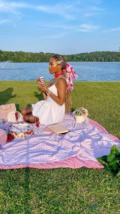 a woman sitting on top of a blanket next to a lake eating cake and drinking tea