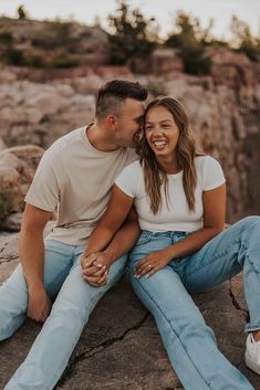 a man and woman sitting on top of a rock near the ocean smiling at each other