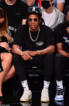 the rapper is sitting down at a basketball game wearing sunglasses and a black hat with his hand on his hip