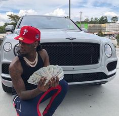 a man sitting on the ground with money in front of a silver bentley - benz