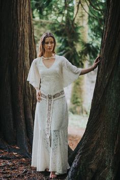 a woman in a white dress standing next to a tree with her hands on the trunk