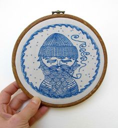 a hand holding a blue and white embroidery on a wooden hoop with a drawing of a bearded man wearing a knitted hat