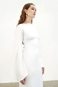 a woman wearing a white dress with bell sleeves and gold hoop earrings standing in front of a wall