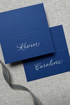 two blue cards with white ink on them sitting next to a silver ribbon and some other items