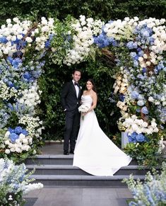 a bride and groom standing in front of an archway with blue and white flowers on it