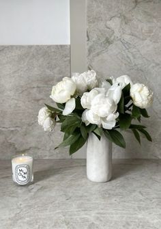 white flowers in a vase next to a candle on a marble counter top with a mirror behind it