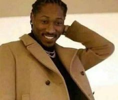 a man with dreadlocks wearing a tan coat and black shirt smiles at the camera