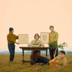 four people standing and sitting around a table in the middle of a field with pictures on it