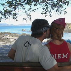 a man and woman sitting on a bench next to each other under a tree near the ocean