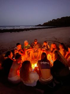 a group of people sitting around a fire pit on the beach at night with lights